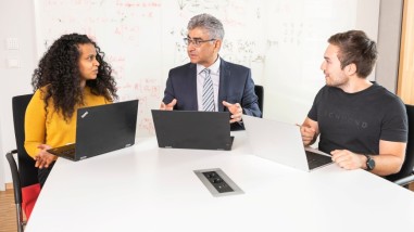 Prof. Ahmad Reza Sadeghi (mitte) sitzt mit einer jungen Wissenschaftlerin und einem jungen Wissenschaftler an einem Tisch und unterhält sich. 3 aufgeklappte Laptops stehen vor ihnen.