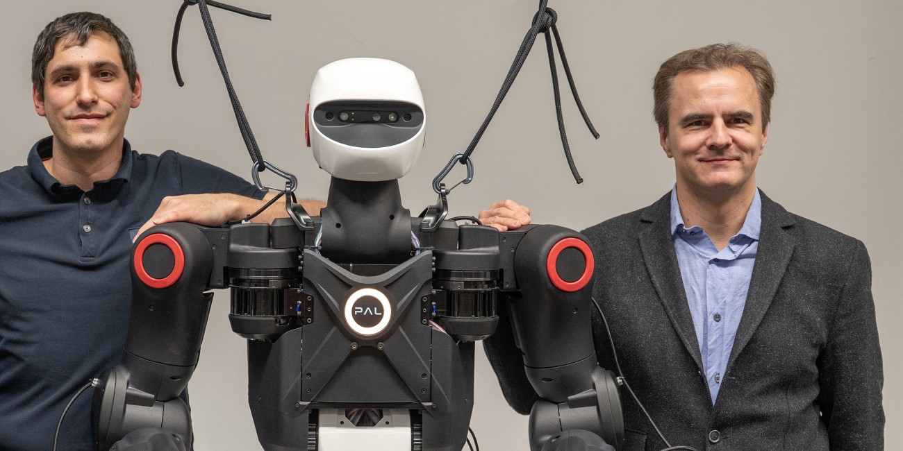 Dr. Oleg Arenz and Professor Jan Peters from the IAS Group at TU Darmstadt standing next to a PAL Robotics humanoid robot, with their hands resting on its shoulders.
