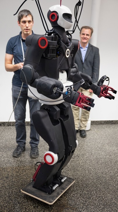 The PAL Robotics humanoid robot in an upright standing position, supported by a harness, with Oleg Arenz and Professor Jan Peters observing from behind.