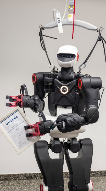 The humanoid robot holding a certificate labeled "Urkunde" in one hand while suspended in a harness.