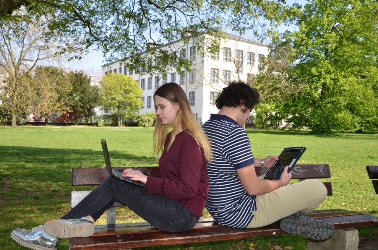 Zwei Studierende mit Laptop auf Schoß Rücken an Rücken vor Piloty-Gebäude